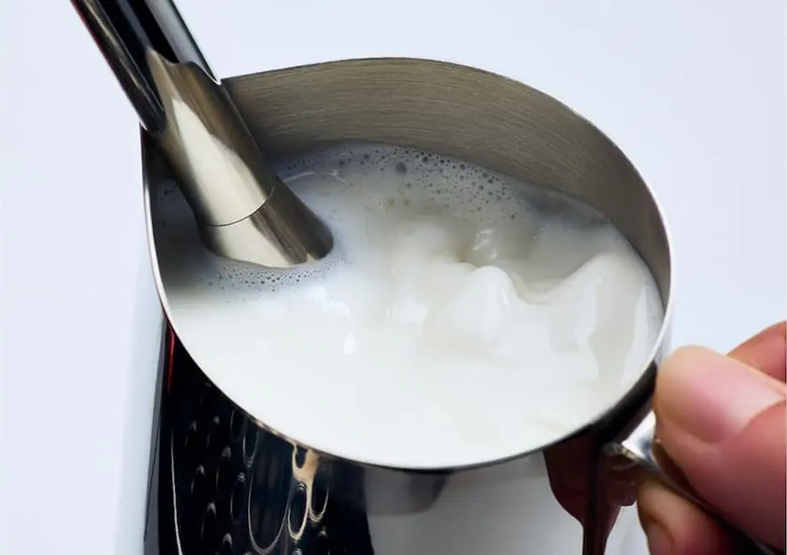 A close-up of a hand holding a metal pitcher filled with milk, being frothed by the steam wand of an Espresso Machine, creating foam on the surface. The background is plain and light-colored.