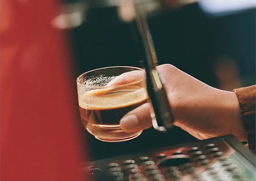 A hand holds a small glass cup filled with espresso under the Fellow Series 1 Espresso Machine, ready to catch the freshly brewed coffee.