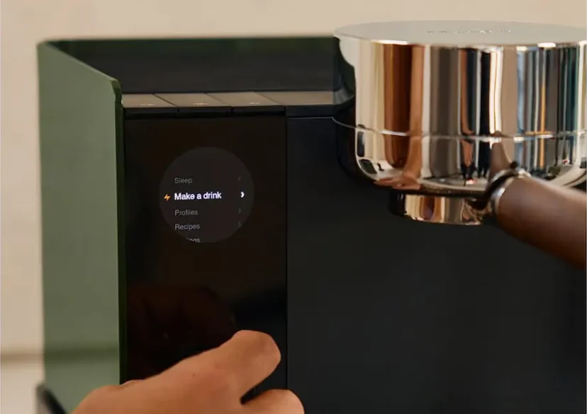 A hand presses a button on the sleek, modern Fellow Series 1 Espresso Machine, its digital display glowing with the option Make a drink.