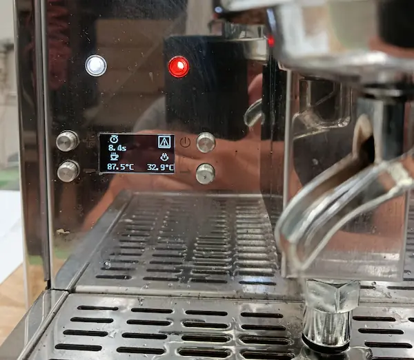 A close-up of an espresso machine in use, showing a digital display with time, temperature, and other settings. The machines portafilter and drip tray are also visible.